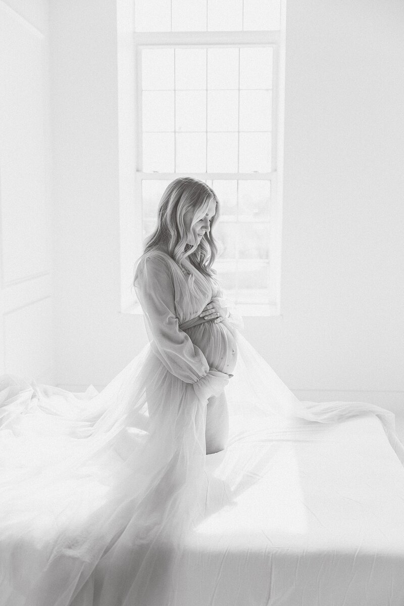 A pregnant woman, captured by a talented Charlotte maternity photographer, is elegantly seated on a bed in a captivating black and white photo.