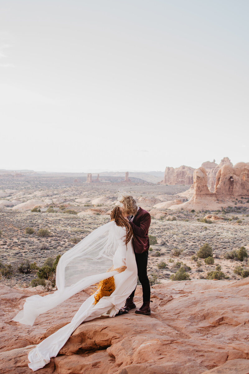 arches national park wedding  with bride and groom on a red rock cliff and kissing with the wind blowing the brides veil and a stunning view of Moab's valley in the distance