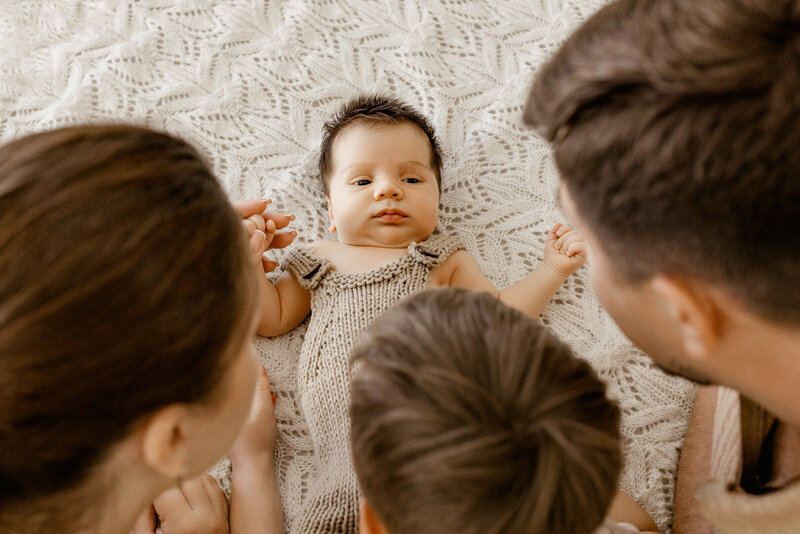 Top view of baby boy in neutral knit romper lying on a textured blanket, surrounded by family gazes during lifestyle newborn photography in Las Vegas