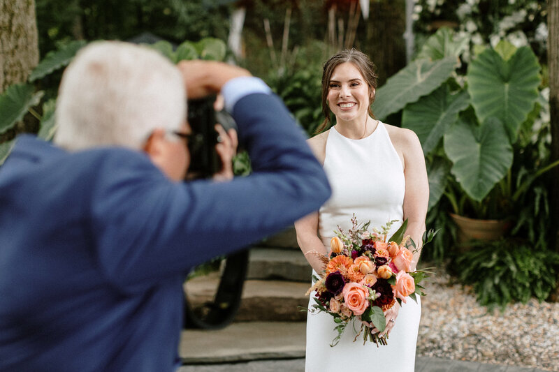 A bride in a white gown poses with a colorful bouquet while a family member photographs the moment in a lush outdoor setting.