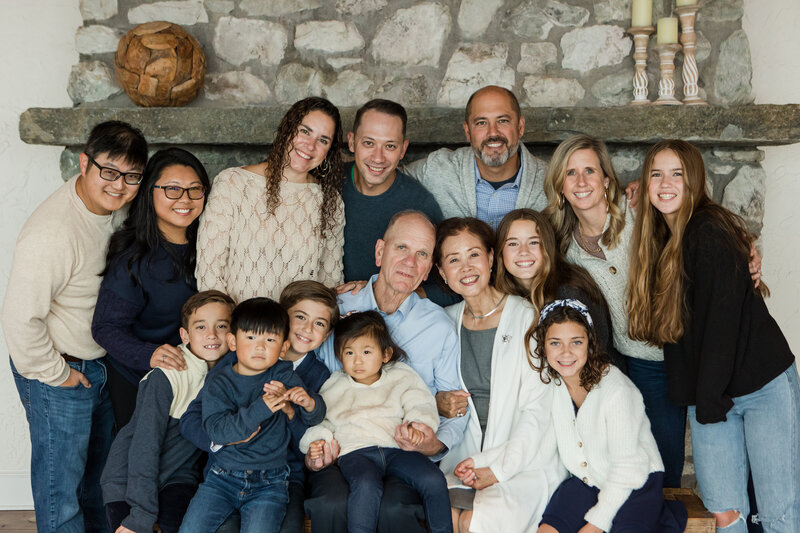 A large family of fifteen leans in and smiles together in front of a stone fireplace all wearing blue, white and cream color coordinated outfits. The grandmother and grandfather are surrounded by their children and grandchildren.