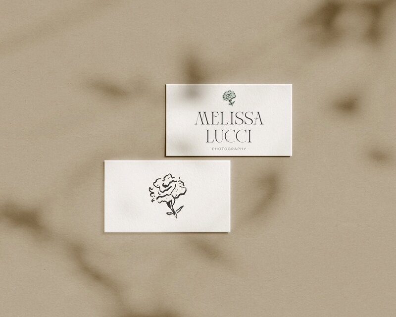 Two business cards on a beige surface. The top card reads "Melissa Lucci Photography" with a flower illustration, crafted by an expert in branding and website design for creatives. The bottom card has only the flower illustration. Shadows are cast across the cards, creating a stylish composition.