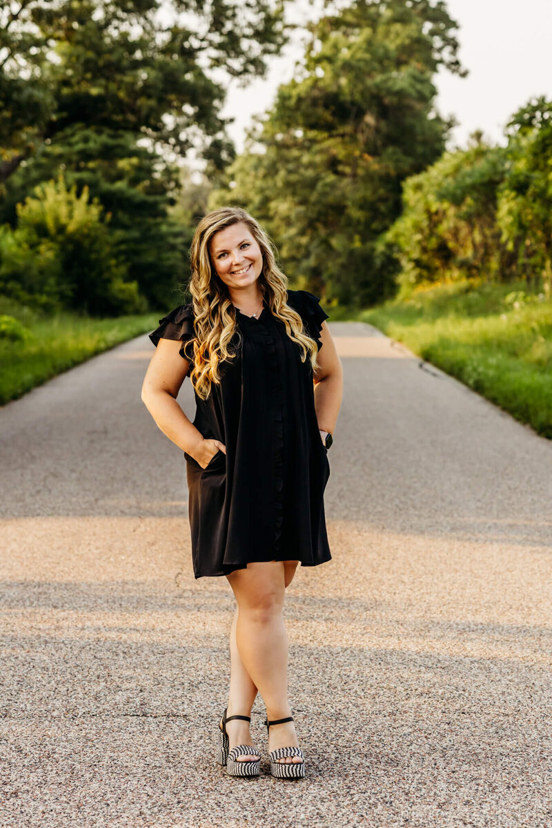 family photographer Ashley Kalbus standing on a back road in a short black dress and chunky heels