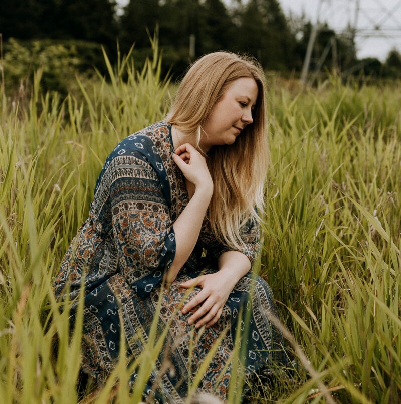 Jaymie Sutherland, Spiritual Healer, sitting on a bench, hand tipping her hat, smiling softly at the camera.