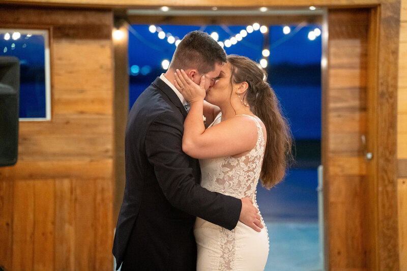 Bride and groom share a romantic kiss inside the barn at Cousiac Manor, with string lights glowing behind them during their wedding reception.