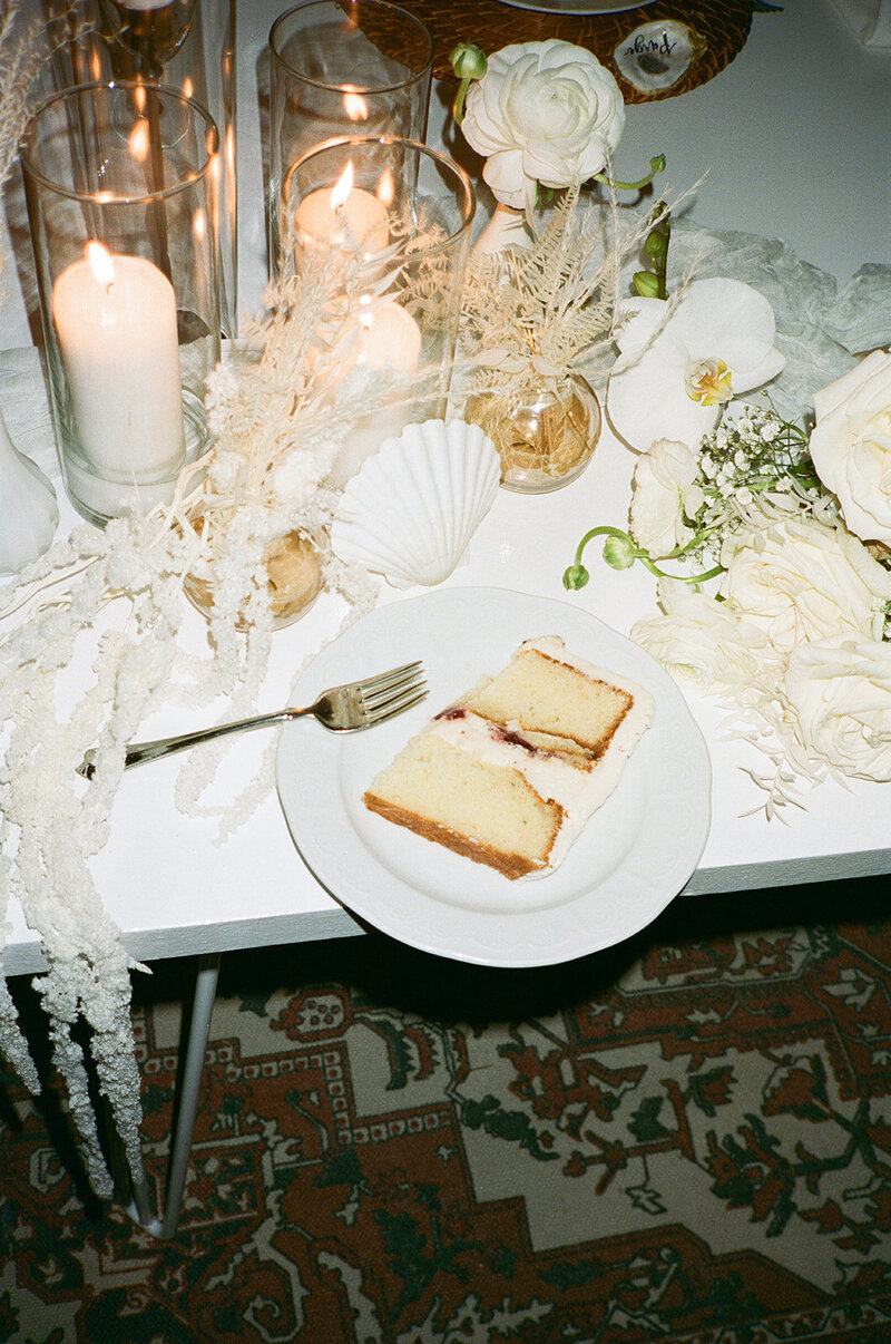 Two slices of wedding cake on a white plate beside an elegant tablescape with candles and white florals, styled by The Eventful Gals.