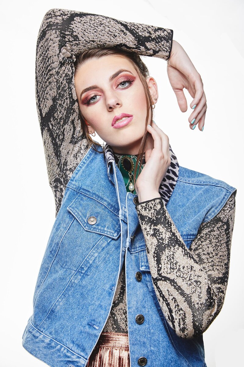 High school model striking a strong editorial pose in a snakeskin print top and denim vest with bold glam makeup – photographed in a white studio during a styled mentorship shoot.