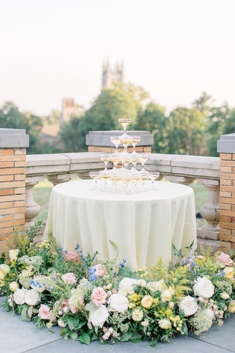 Outdoor shot of a champagne tower on a table surrounded by floral arrangements
