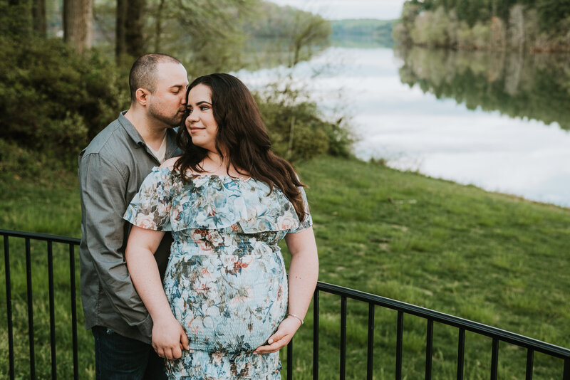 Dad in gray and black standing next to pregnant wife in blue floral dress holding belly in Montgomery County Maryland by a beautiful river.
