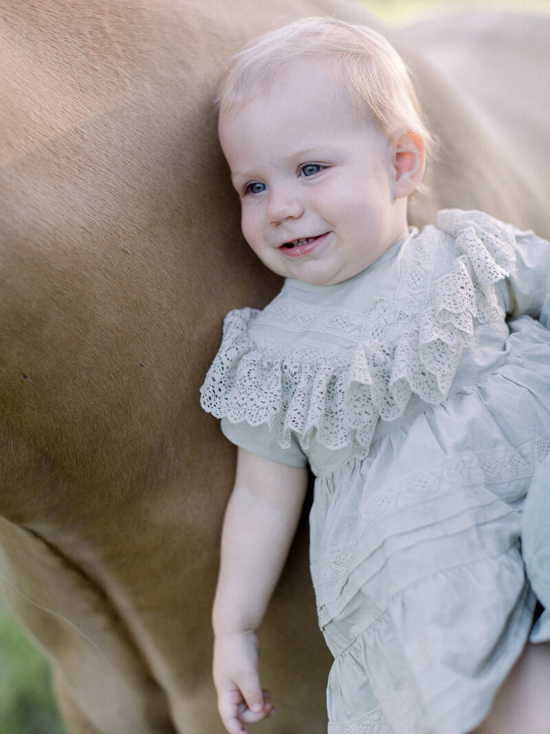 Toddler girl snuggles her horse while smiling in a green ruffled dress on a horse farm by NH family photographer Fieldstone Studio.
