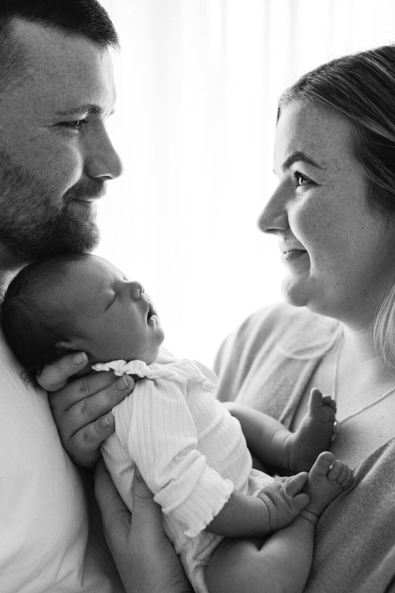 Parents look at eachother and smile whilst holding thier baby