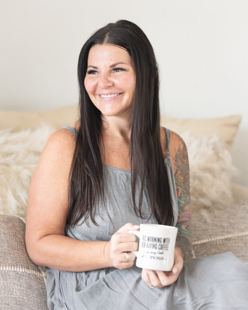 portrait of owner in grey jumper with beige pillows in the back drinking coffee looking out window smiling