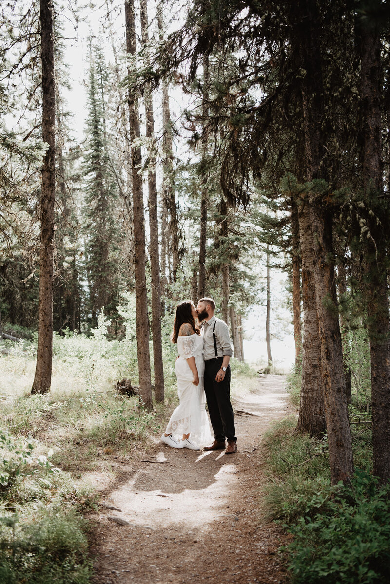 jackson hole wedding photos with bride and groom kissing as they walk a trail through the woods next to a beach with tall thin trees surrounding them
