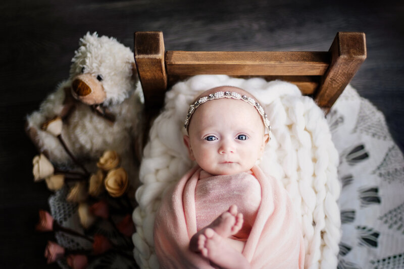 Awake and alert, this newborn girl’s portrait features a cozy pink wrap, floral touches, and a plush teddy friend.