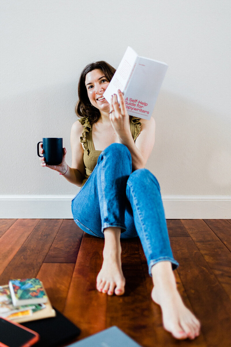 Photo of Andrea Shah, a copywriter for wedding pros and educators, sitting on the floor while holding a book and a mug