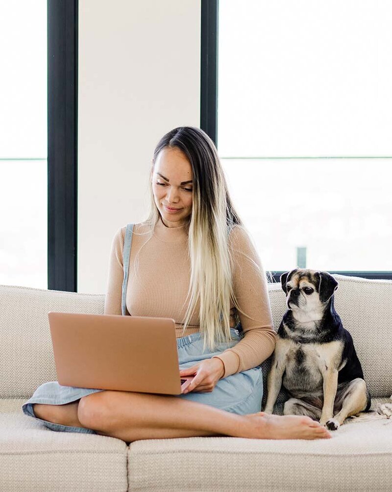 Young Asian female entrepreneur sitting on a cozy white couch with her pug, both looking at her pink MacBook, representing trust in Xanthe Bookkeeping’s business values.