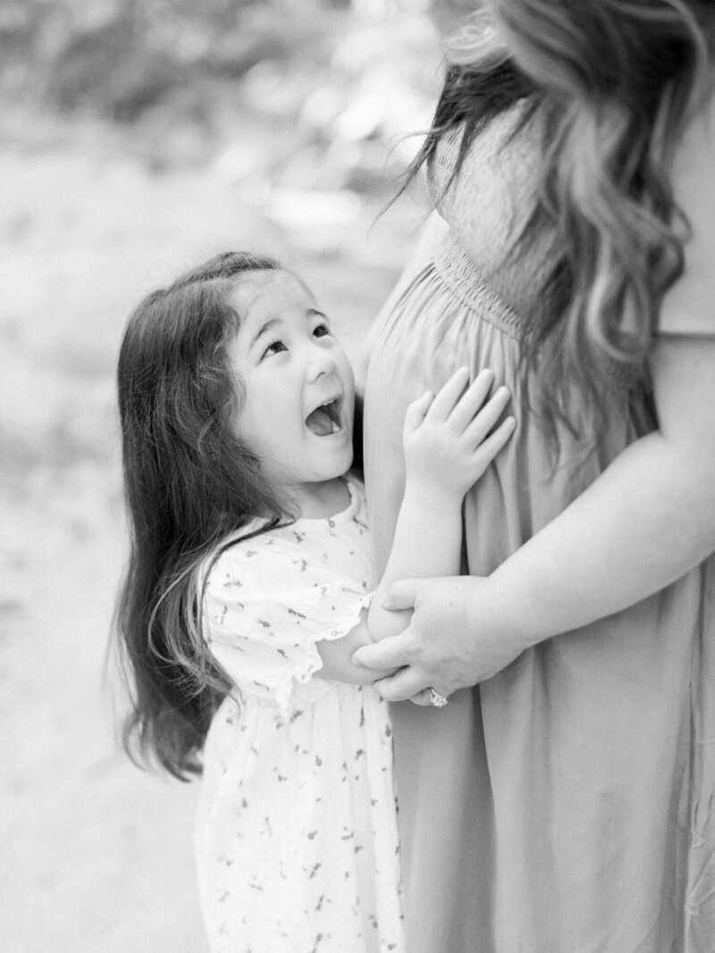 A joyful young girl in a light dress smiles up at a pregnant woman's belly, gently touching it. They stand close, sharing a tender, happy moment.