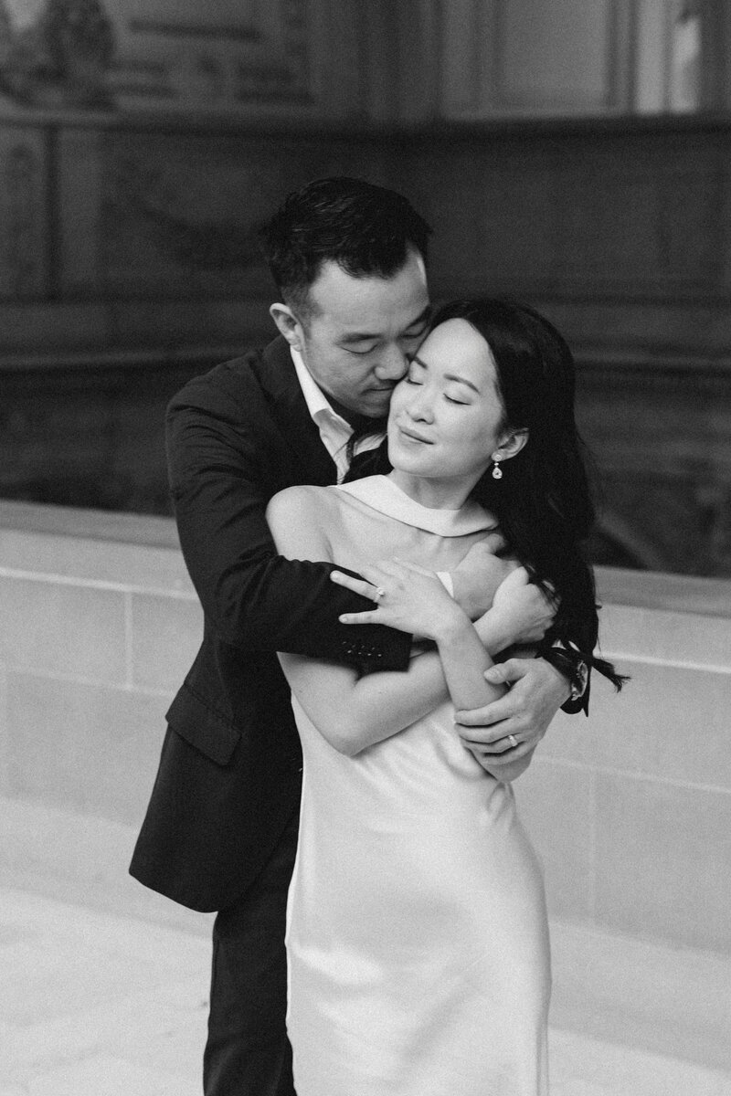 A monochrome photoshoot of a man in a suit kissing a woman in a white dress on the forehead as they embrace tenderly during their elopement photoshoot in San Francisco City Hall