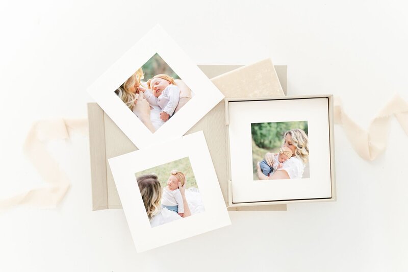 flatlay photo of a matted print box on a white background. The box has a tan, linen cover. Three matted prints are laid on top of it showing photos of an outdoor newborn session.