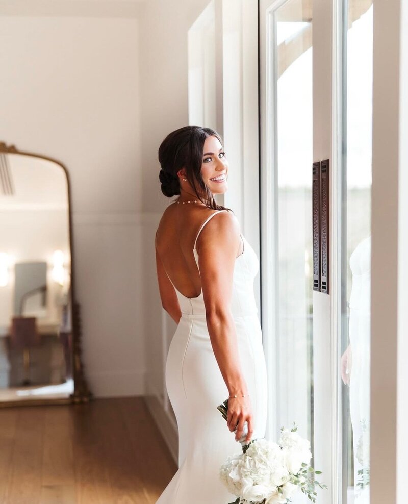 A bride in a white dress, holding flowers, smiles by a window with reflections, encapsulating the elegant room setting. Her joy is heightened by her amazing bridal beauty experience in Texas, perfectly complementing her radiant wedding day glow.