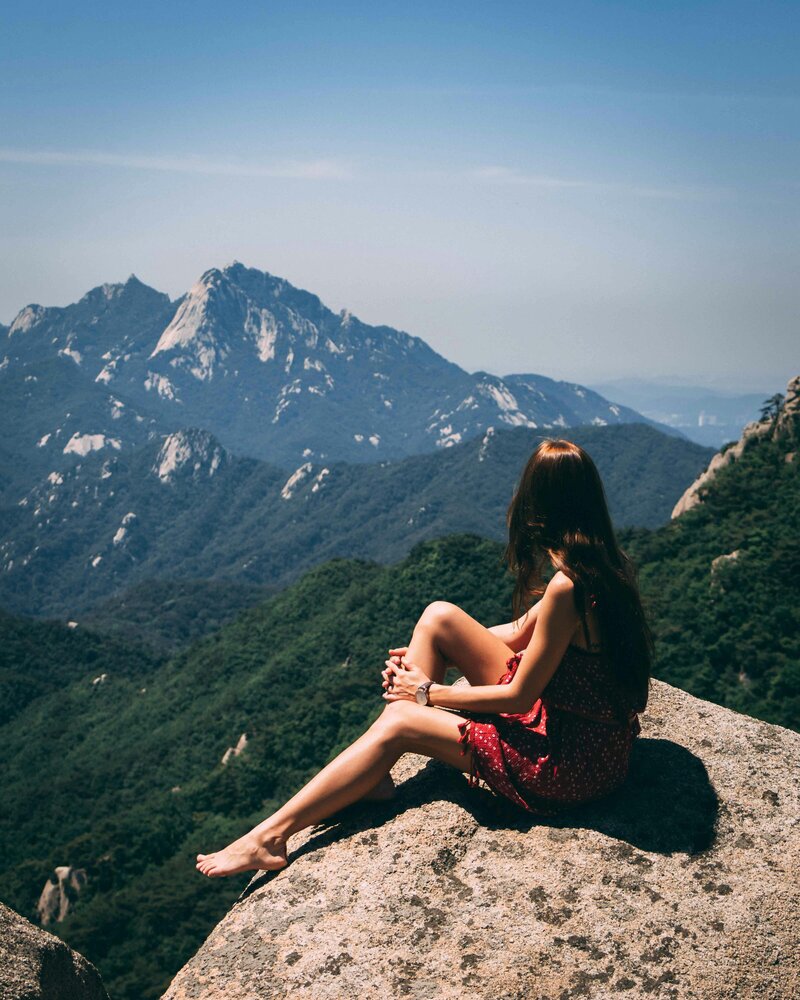 Image of a young femme presenting person sitting barefoot in a red dress on a mountain top