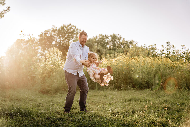 A father joyfully swings his young daughter in the air by her arms in a grassy field during golden hour, with a background of yellow wildflowers.