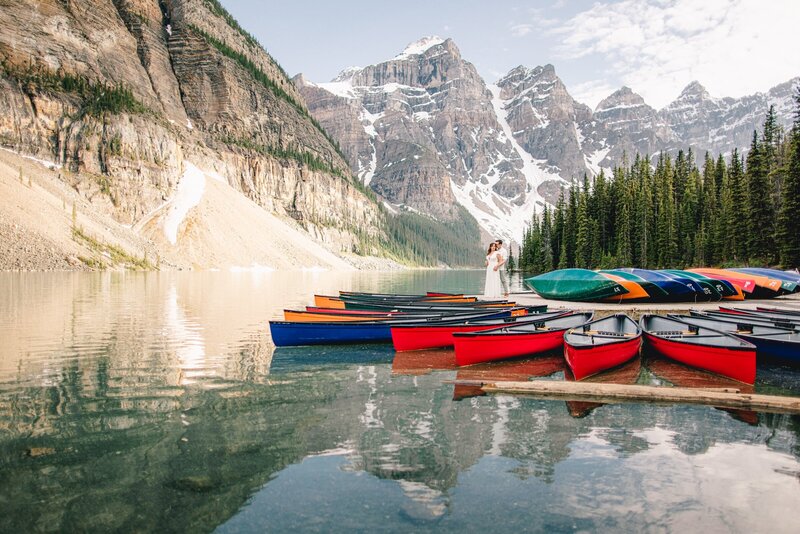 An evening sunset engagement photo captures a couple near the Moraine Lake boat dock, with canoes and the stunning Valley of the Ten Peaks in the background. The warm sunset light enhances the serene and romantic atmosphere at this iconic location.