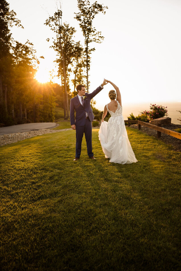 A groom twirls his bride on the grass during sunset.