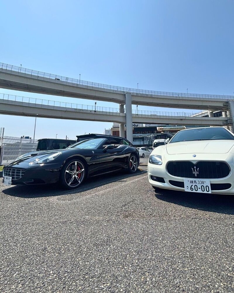 A black Ferrari California and a white Maserati Quattroporte parked side by side under the Tokyo expressway at Daikoku Parking Area, ready for a luxury drive experience.