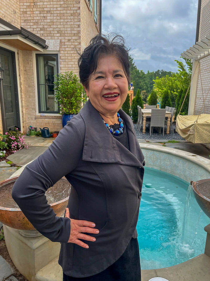 Portrait of author and speaker, Nita Tin, smiling warmly in a gray dress jacket and black dress with hand on hip, standing outdoors on her patio with flowers and pool in the background, reflecting her present-day life in Georgia.