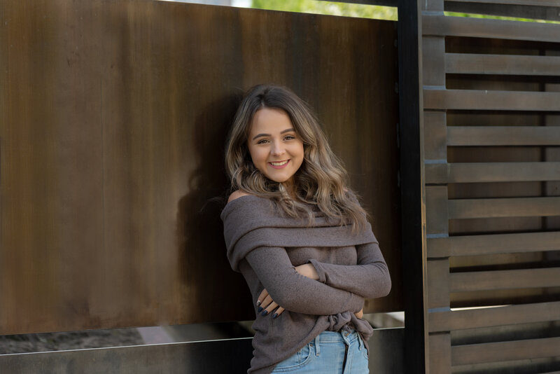 High school senior girl smiling and wearing brown sweater leaning against a brown wall