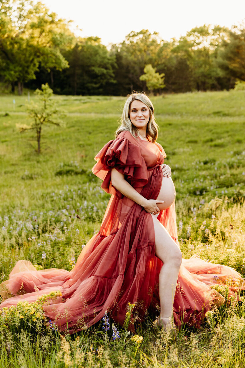 gorgeous mom to be wearing large gown that shows her baby bump while standing in a flower field at sunset near Green Bay