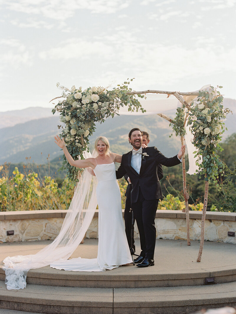 couple under their huppah after they have said "I do" at Holman Ranch with vineyard behind them and huge smiles