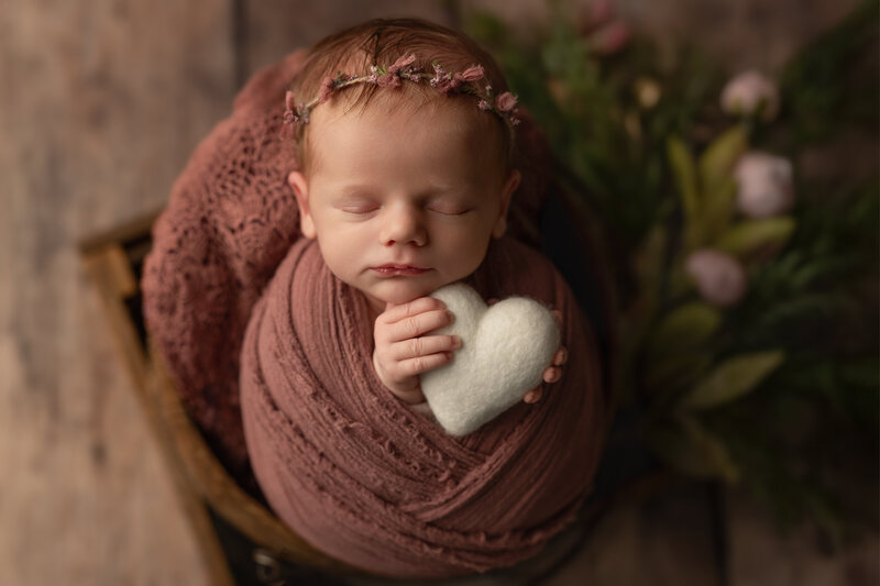 Newborn girl posed in a mauve wrap with a white heart and floral crown – rustic, heartfelt newborn portrait.