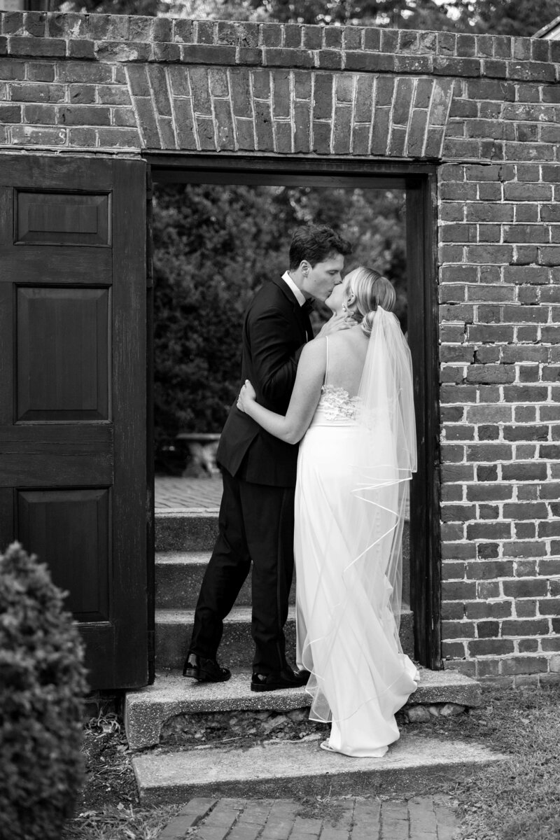 A bride and groom share a kiss on the steps of a brick building. The bride wears a strapless gown and veil, while the groom is in a suit. The setting suggests an intimate, romantic moment. Black and white photo.