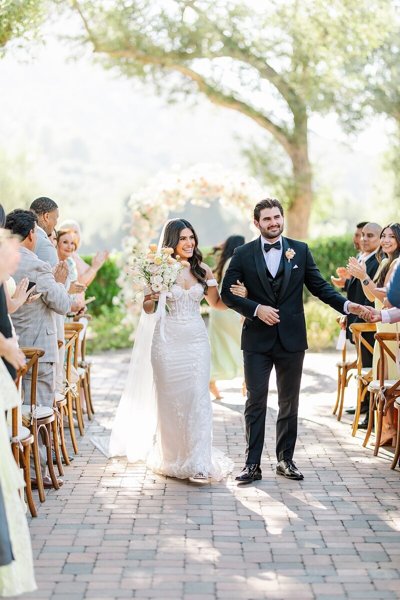 Bride and groom walking hand in hand during their wedding ceremony recessional, guests clapping as they glow with excitement at Mt. Woodsen Castle in Ramona.