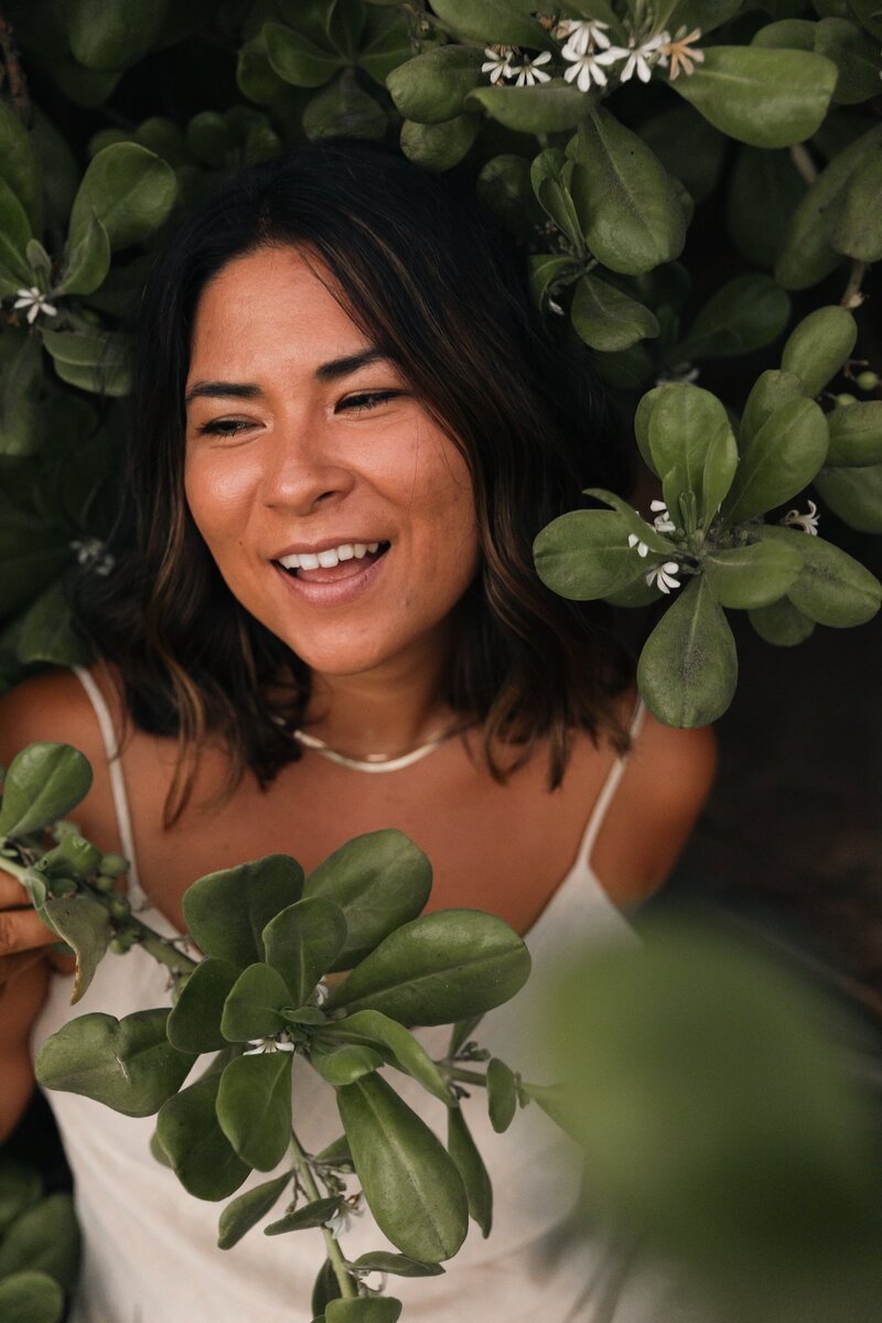 Amber, surrounded by tropical greenery, smiles while looking off camera to the left mid laugh.