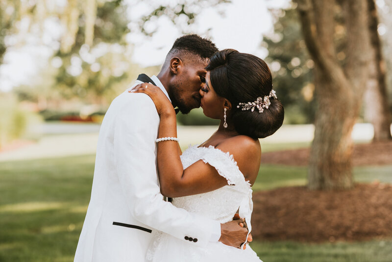 A bride and groom share a tender kiss in a lush park setting. The bride, wearing a white gown with feathered details and a pearl bracelet, has her hair elegantly styled in a bun adorned with a floral hairpiece. The groom, in a white suit with black accents, gently holds her. The background features soft greenery and trees, creating a serene and romantic atmosphere.