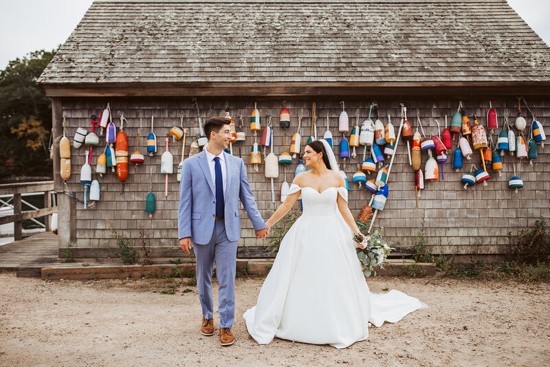 a wedding in york maine with the couple walking toward the camera smiling at each other holding hands with boueys behind them.