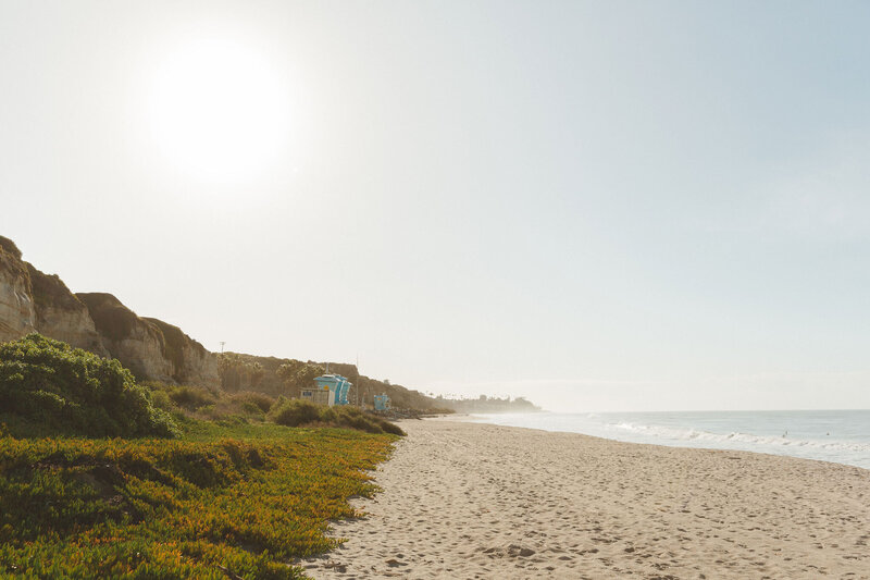 A sunny beach in San Clemente, California, used for a Shindig Social styled shoot. The image captures the clear sky, sandy shore, ocean waves, and coastal cliffs with greenery, highlighting the serene and natural beauty of the location.