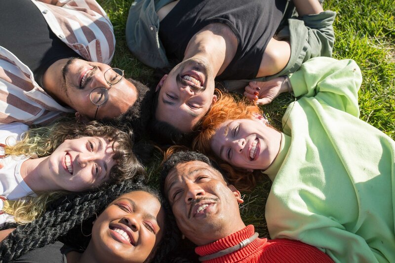 This image shows six young people with diverse racial and gender presentations. All six are lying in the grass, with their heads together to form a circle, as they smile and laugh.
