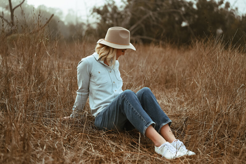Emily Collins sitting in a dry grassy field wearing a hat, light button-up shirt, and jeans, looking down with a relaxed smile.