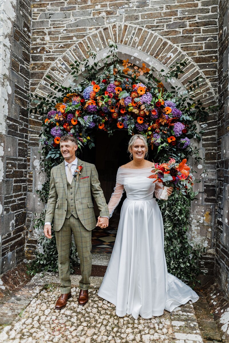 Bride and groom exiting church in Cornwall after getting married with a bright colourful arch around the doorway