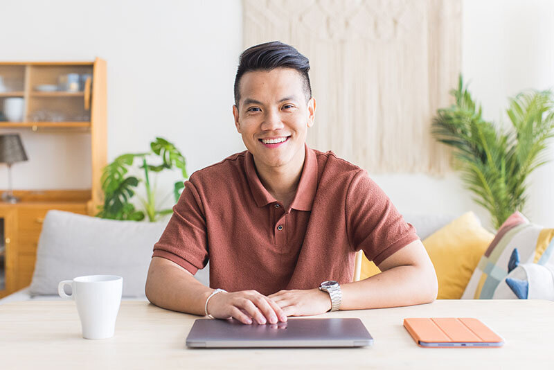Sho Dewan, young Asian man, career coach and founder of Workhap, sitting at a desk with his laptop, tablet and a white coffee mug, looking and smiling directly at the camera