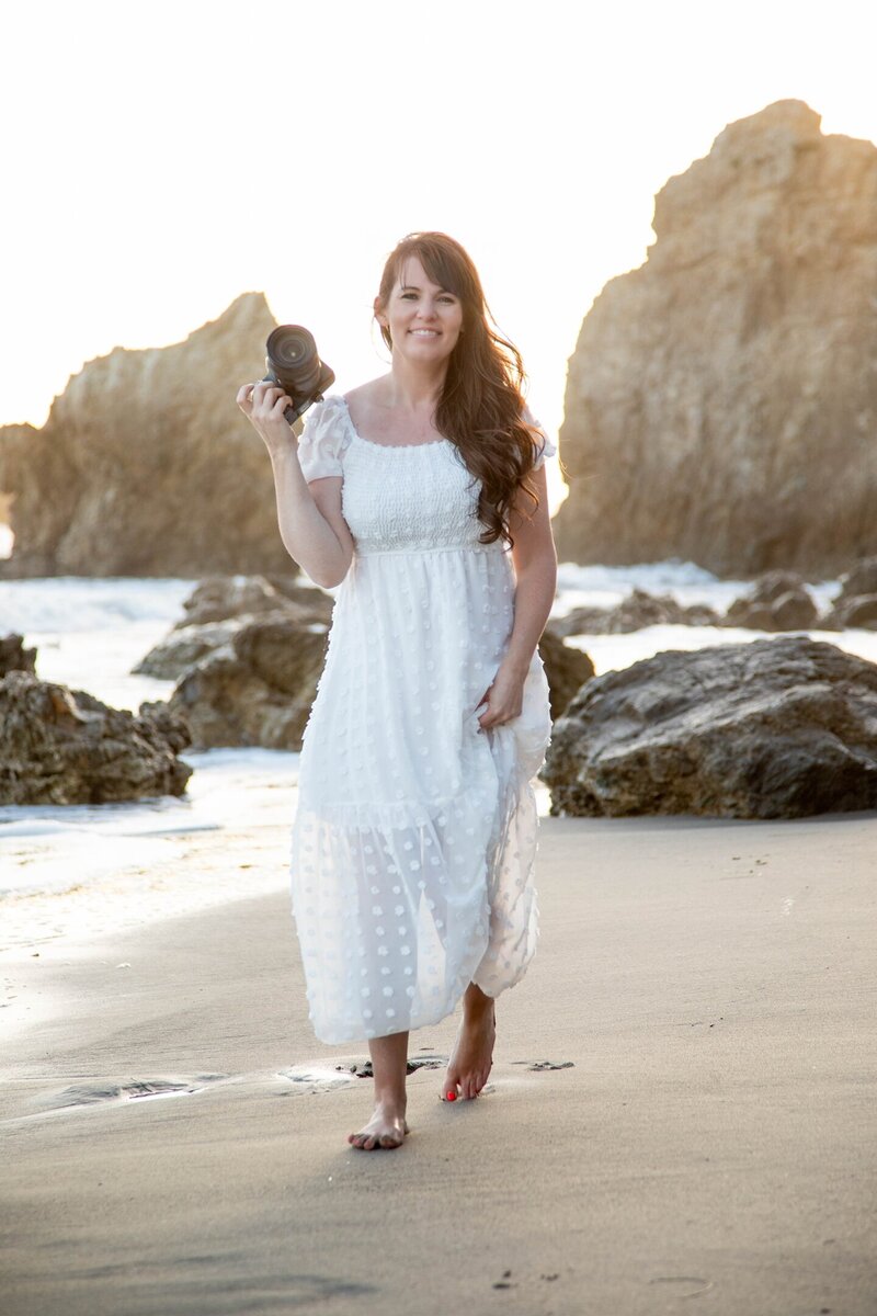 Heidi holding camera with one hand, dress with the other while walking on California beach | Maine Wedding and Portrait Photographer