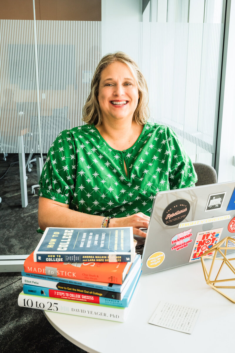 Mya Andersen smiling in a green top sits at a desk with a laptop and a stack of books in a modern office, ready to offer Academic Life Coaching to students.