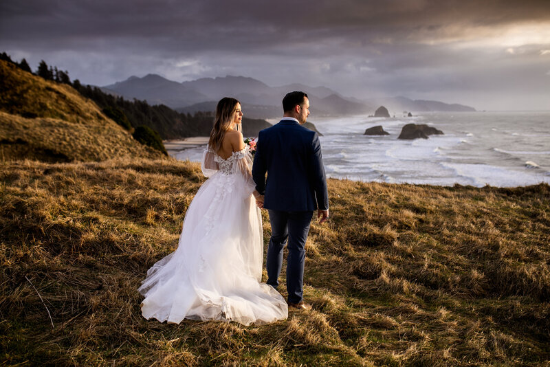 A bride and groom walking hand-in-hand along a coastal cliff in Oregon, with dramatic ocean views, captured by an Oregon elopement photographer.