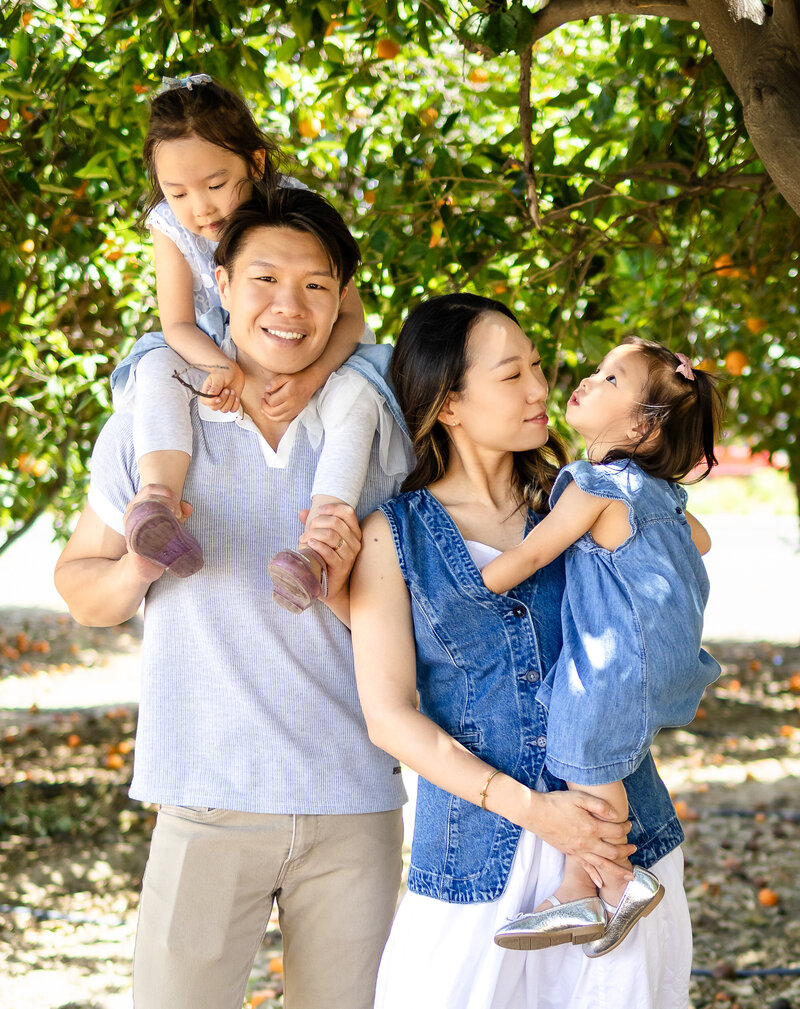 Candid family photo under orange tree in San Mateo featuring parents with two young daughters during natural light lifestyle session by Ellobelle Photography