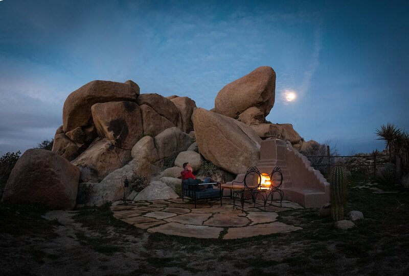 A man sits near a glowing fireplace tucked into rounded pile of rocks during an adventure in Joshua Tree National Park, perfect for adventure luxe destination elopements. 