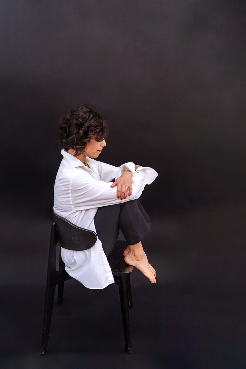 A woman in a white shirt and black pants sitting on a black chair with her arms wrapped around her knees for speaking engagement photos